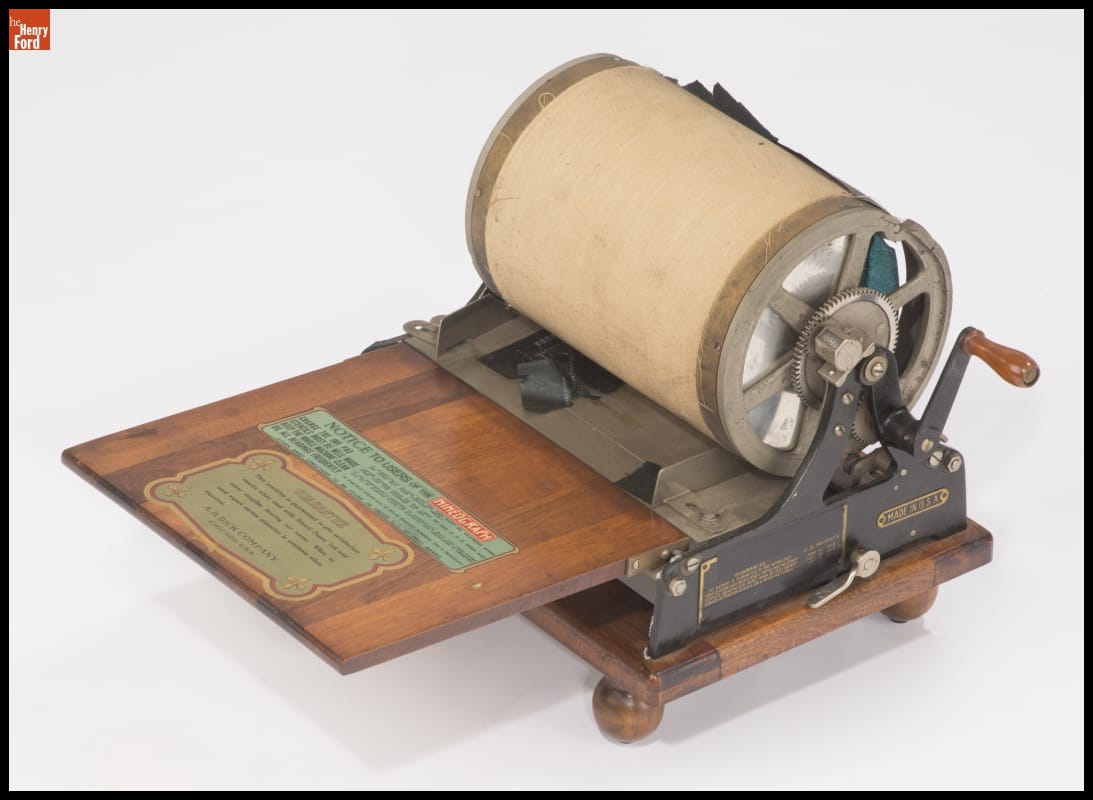 Photograph of a machine that has a flat, wooden bed and a roller with a hand crank.